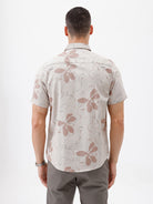 A man wearing a short-sleeve button-up shirt with a beige and brown floral pattern, paired with gray trousers, standing against a plain white background.