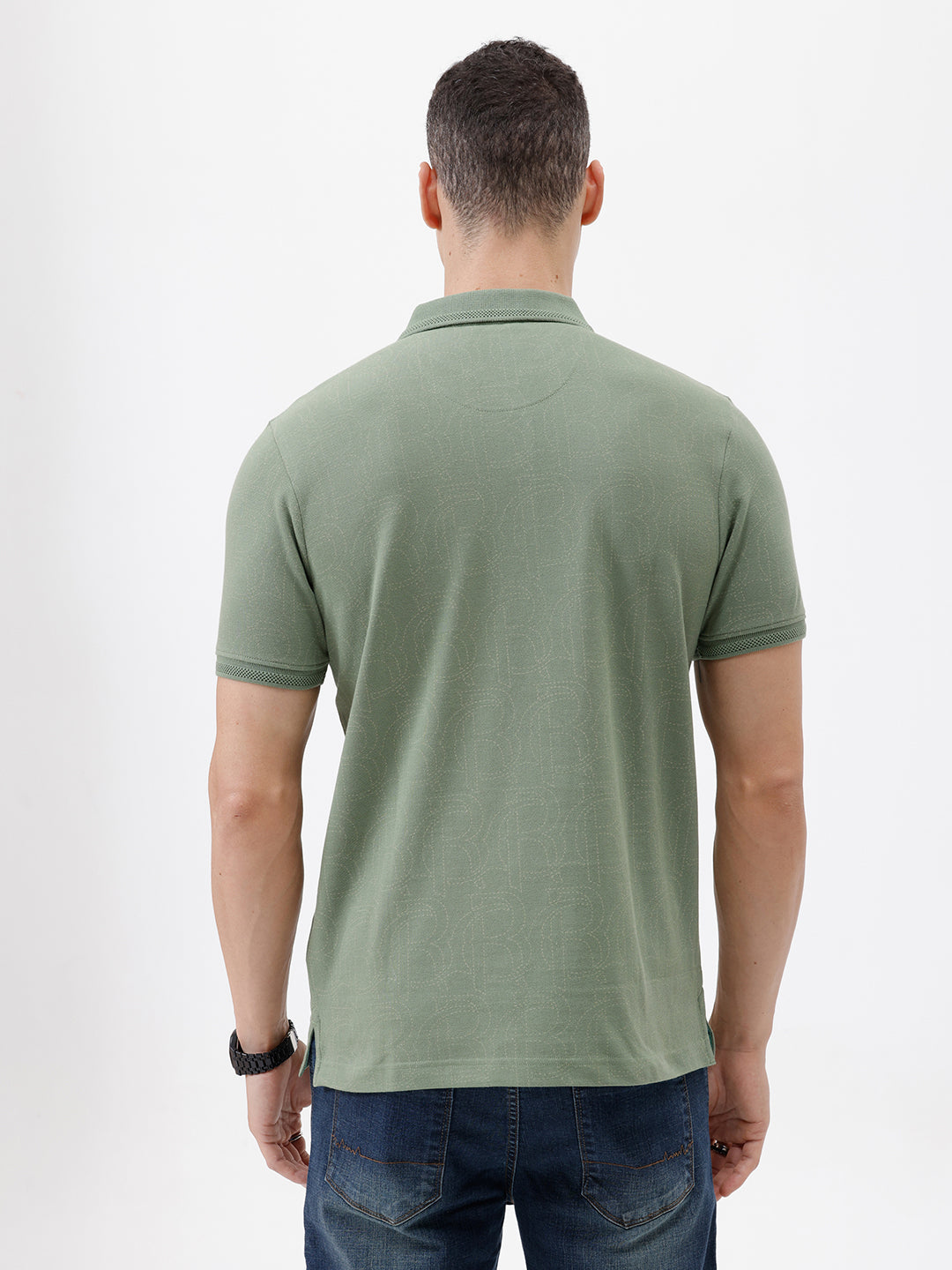 A man wearing a short-sleeve polo shirt in sage green, paired with dark blue jeans, standing against a plain light gray background.