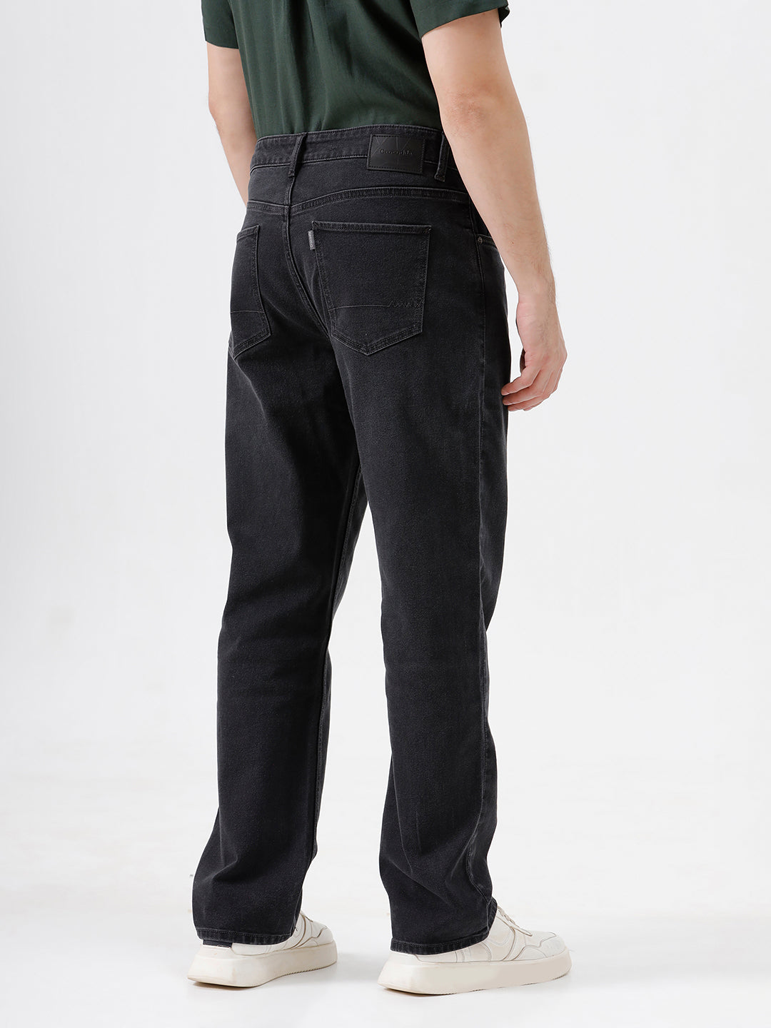 A person wearing dark washed denim jeans, a dark green shirt, and white sneakers, shown from the chest down against a plain light gray studio background.