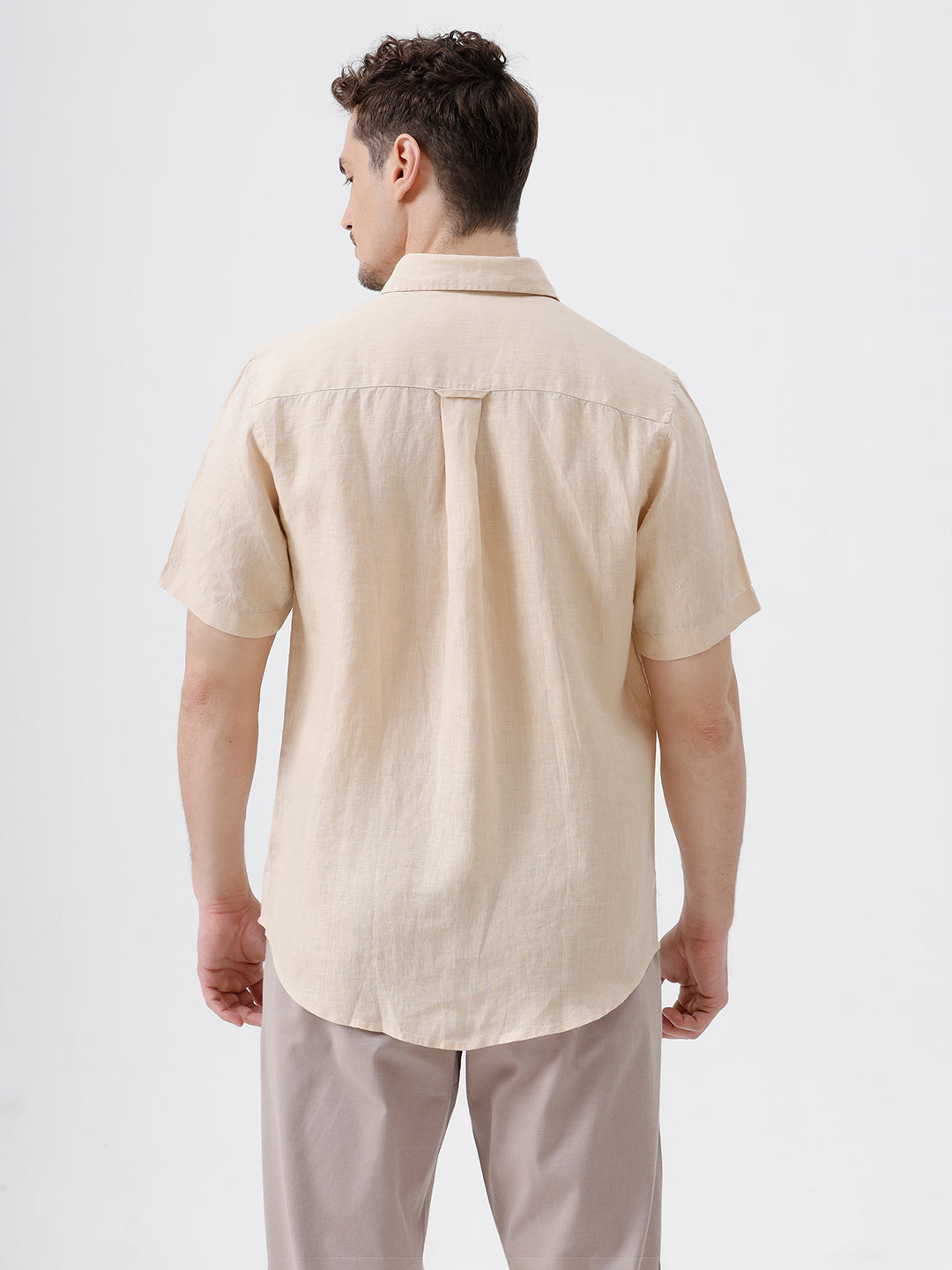 A man wearing a light beige short-sleeve linen button-up shirt and taupe trousers, standing against a plain white studio background.