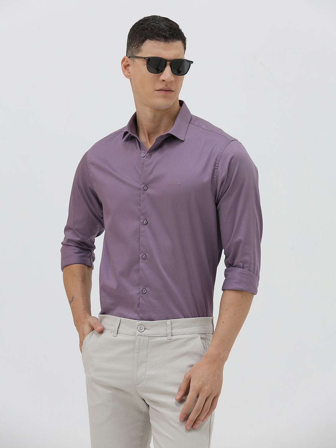 A man wearing a purple long-sleeve button-down shirt with rolled-up sleeves and light beige trousers, standing against a plain light gray background.