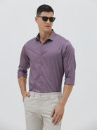 A man wearing a purple long-sleeve button-down shirt with rolled-up sleeves and light beige trousers, standing against a plain light gray background.