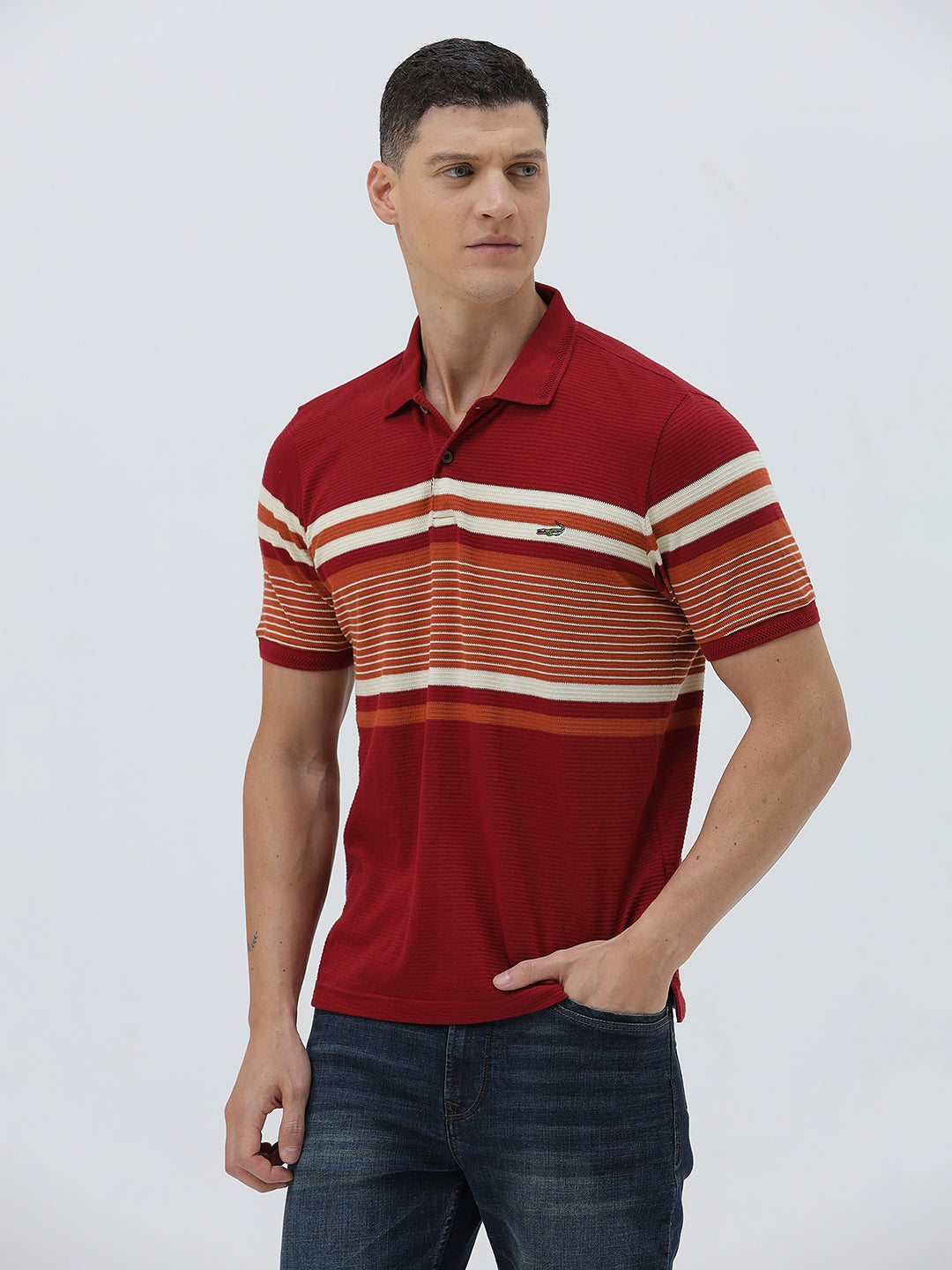A man wearing a red short-sleeve polo shirt with horizontal cream and rust-orange stripes across the chest and sleeves, paired with dark blue jeans and black sunglasses, against a light gray studio background.