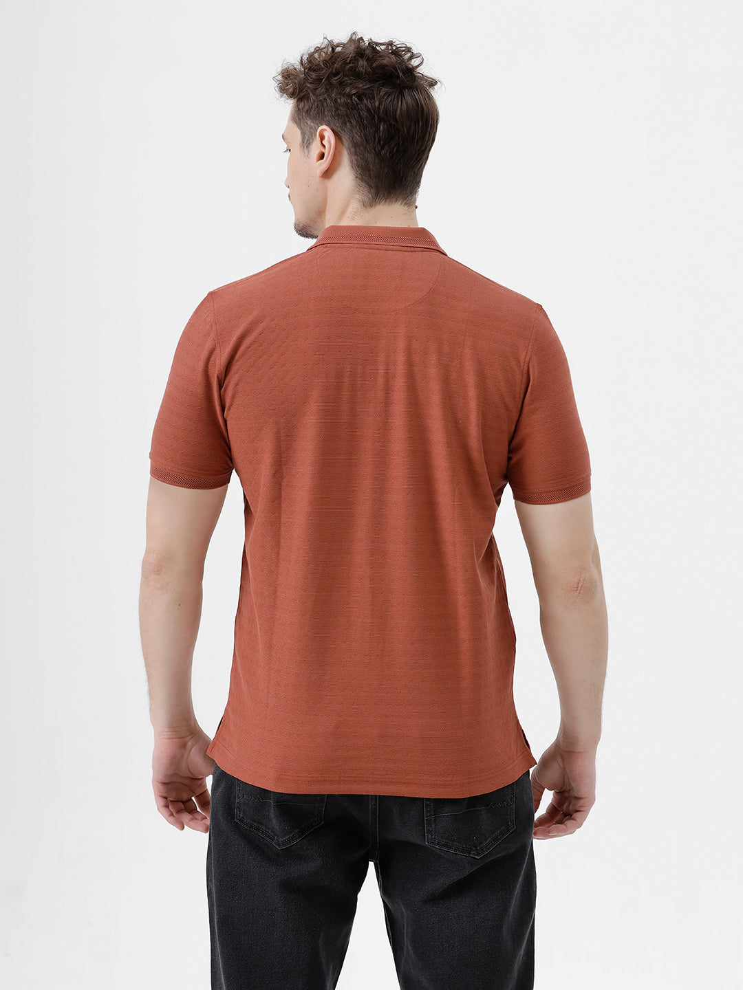 A man wearing a rust-orange short-sleeve polo shirt with a subtle horizontal stripe pattern, paired with dark jeans, standing against a plain white studio background.