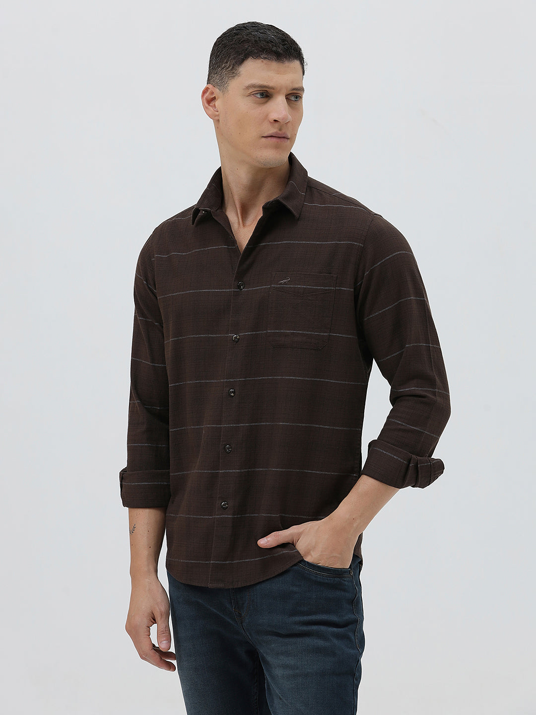 A man wearing a dark brown long-sleeve button-up shirt with thin horizontal stripes, paired with dark blue jeans, standing against a light gray studio background.