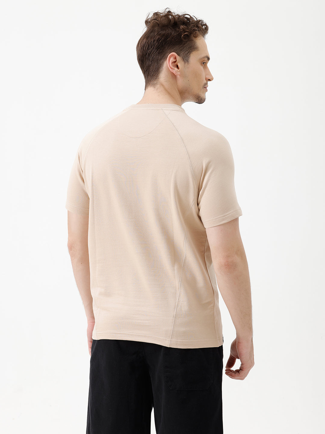 A man wearing a light beige short-sleeve t-shirt with a subtle tonal logo on the chest, paired with black pants, standing against a plain white studio background.