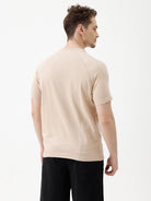 A man wearing a light beige short-sleeve t-shirt with a subtle tonal logo on the chest, paired with black pants, standing against a plain white studio background.