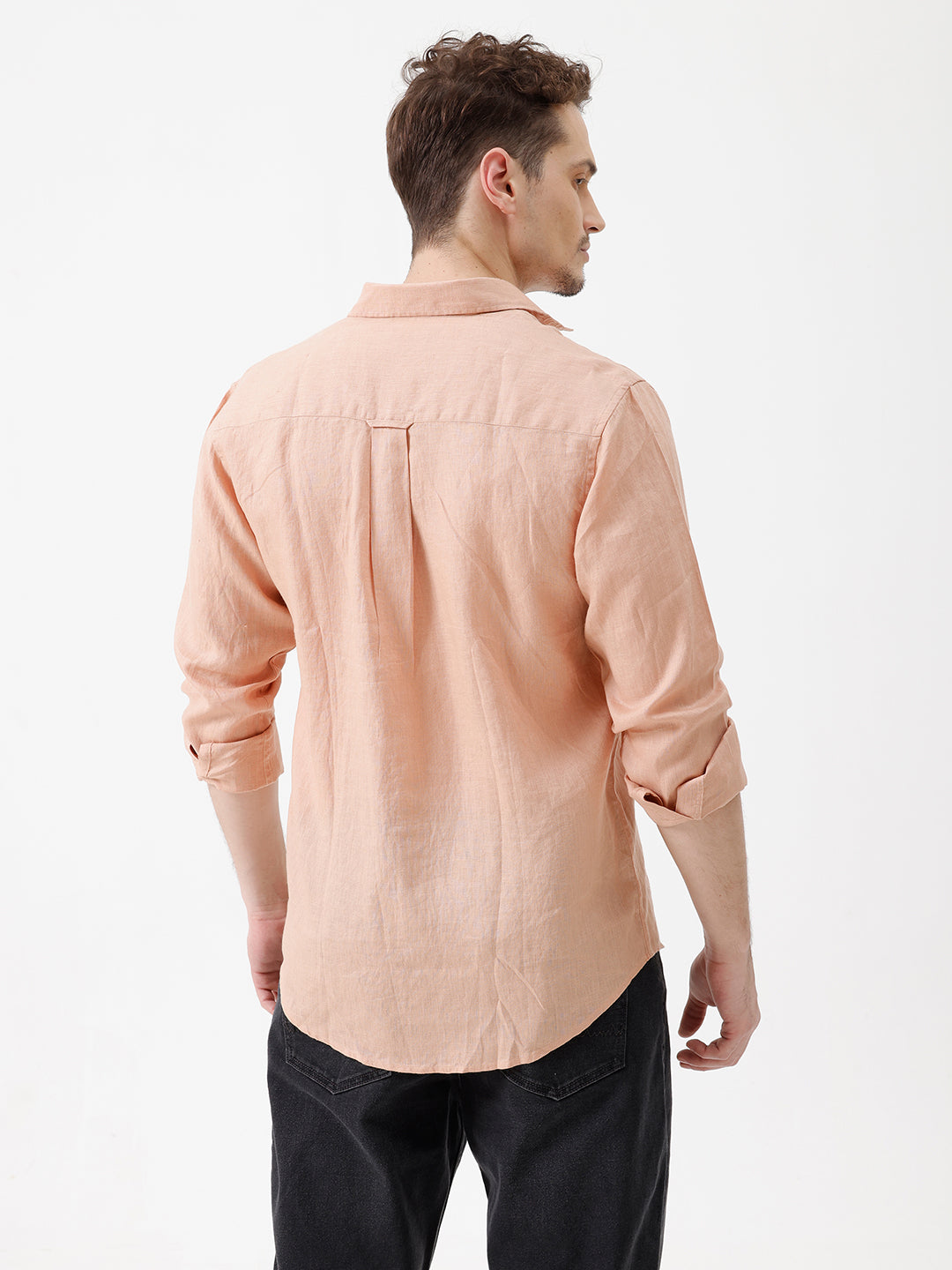 A man wearing a light peach-colored linen button-down shirt with dark jeans, standing against a plain white studio background.