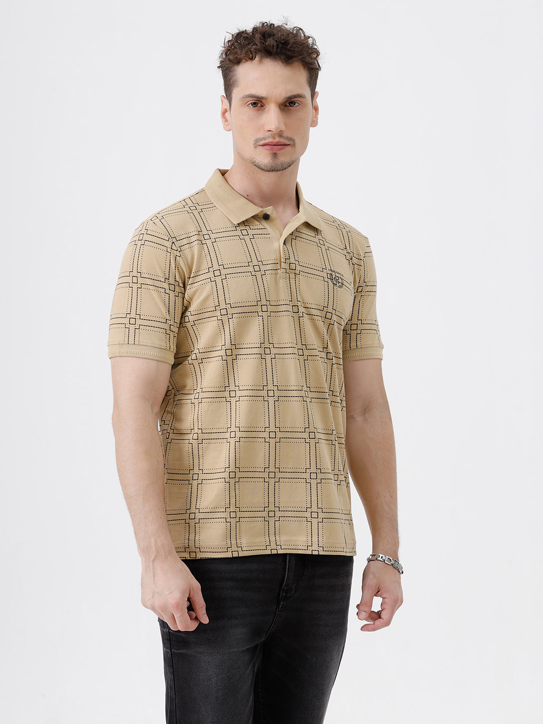 A man wearing a beige short-sleeve polo shirt with a brown grid pattern, paired with black jeans, standing against a plain white studio background.