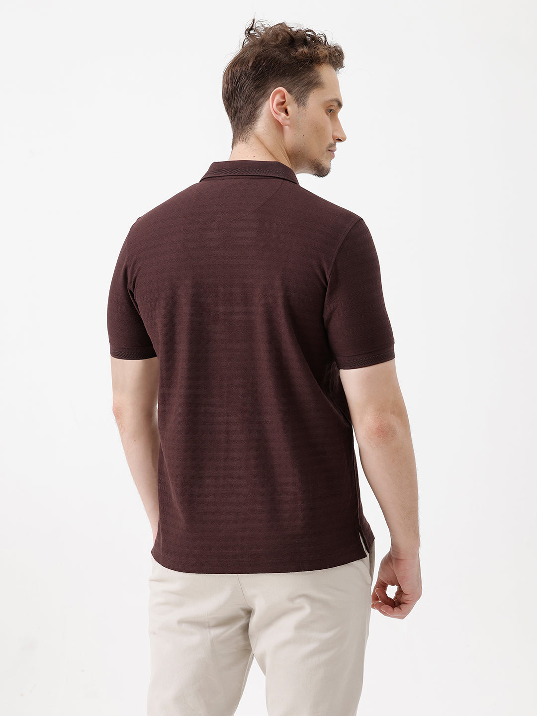A man wearing a dark brown short-sleeve polo shirt with a subtle horizontal stripe pattern and light beige trousers, standing against a plain white studio background.