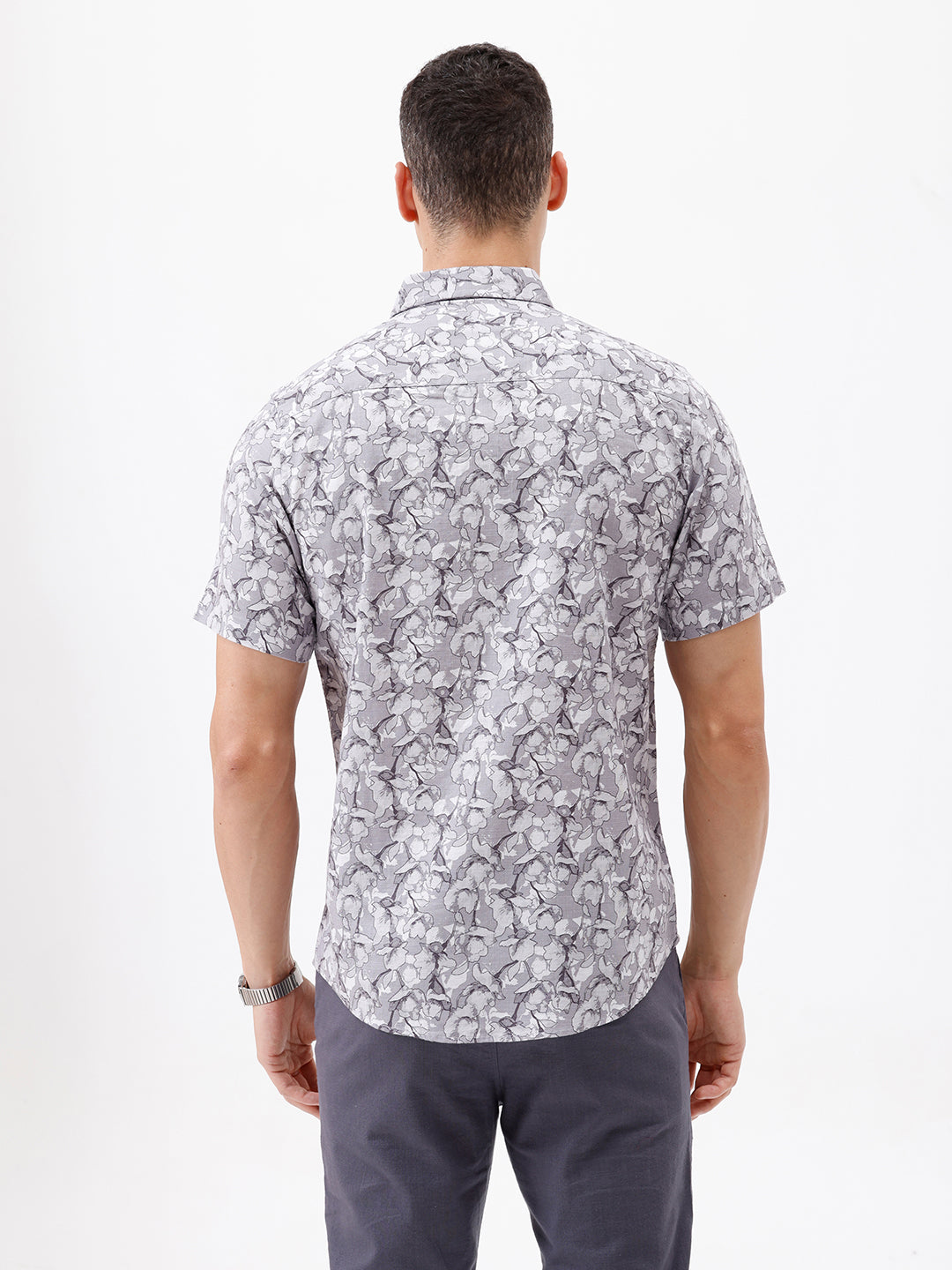 A man wearing a short-sleeve button-up shirt with a gray and white abstract pattern, paired with dark gray trousers, standing against a plain white studio background.
