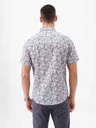 A man wearing a short-sleeve button-up shirt with a gray and white abstract pattern, paired with dark gray trousers, standing against a plain white studio background.