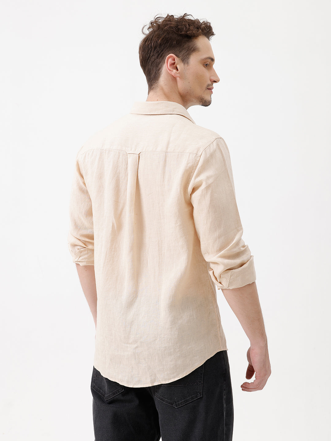 A man wearing a light beige linen button-up shirt with rolled-up sleeves and dark jeans, standing against a plain white studio background.
