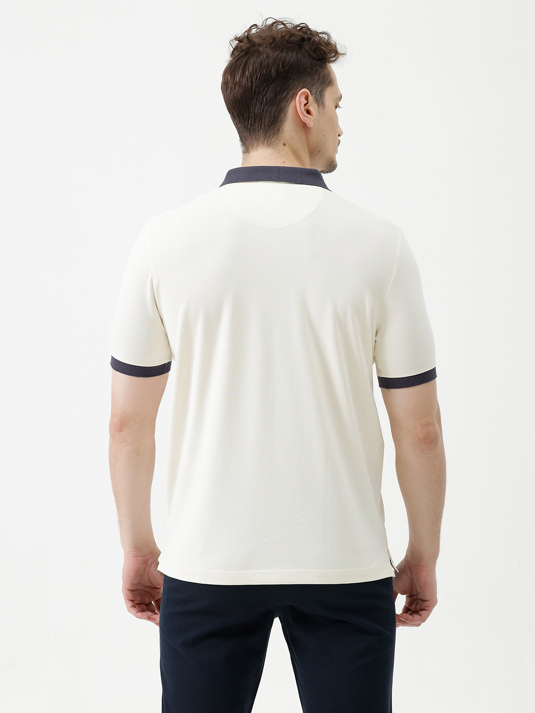 A man wearing a short-sleeve polo shirt in off-white with navy blue and gold trim on the collar and sleeve cuffs, paired with dark navy trousers, against a plain light gray background.