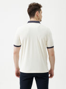 A man wearing a short-sleeve polo shirt in off-white with navy blue and gold trim on the collar and sleeve cuffs, paired with dark navy trousers, against a plain light gray background.