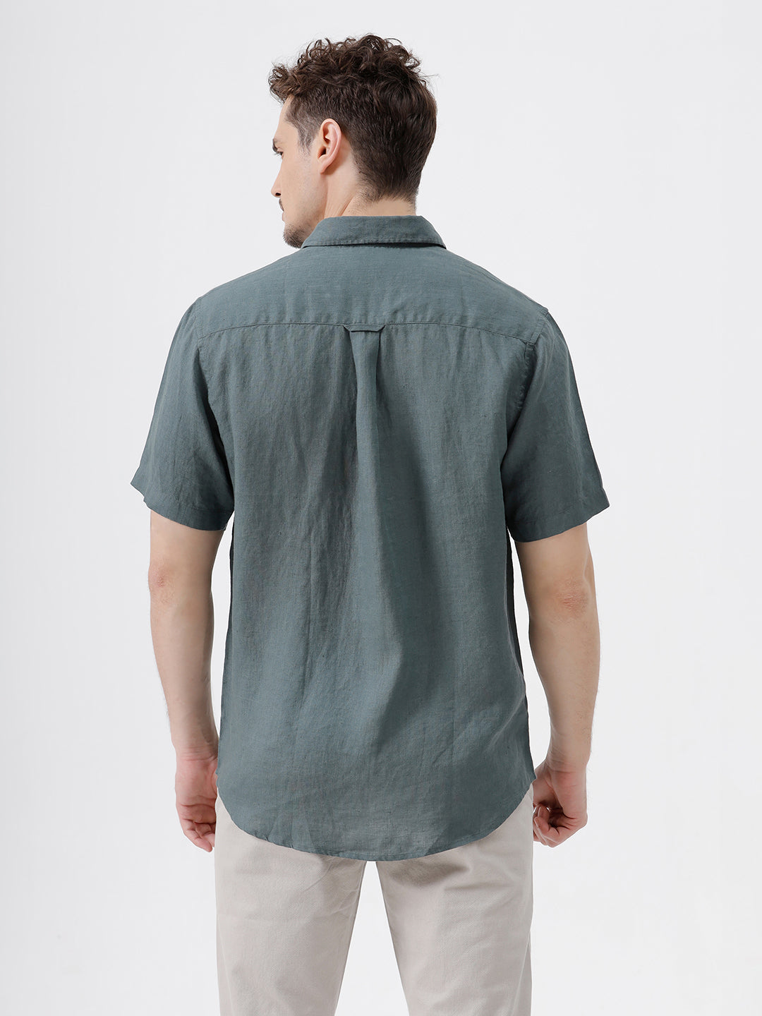 A man wearing a short-sleeve button-up shirt in a muted slate green color, paired with light beige trousers, standing against a plain light gray background.
