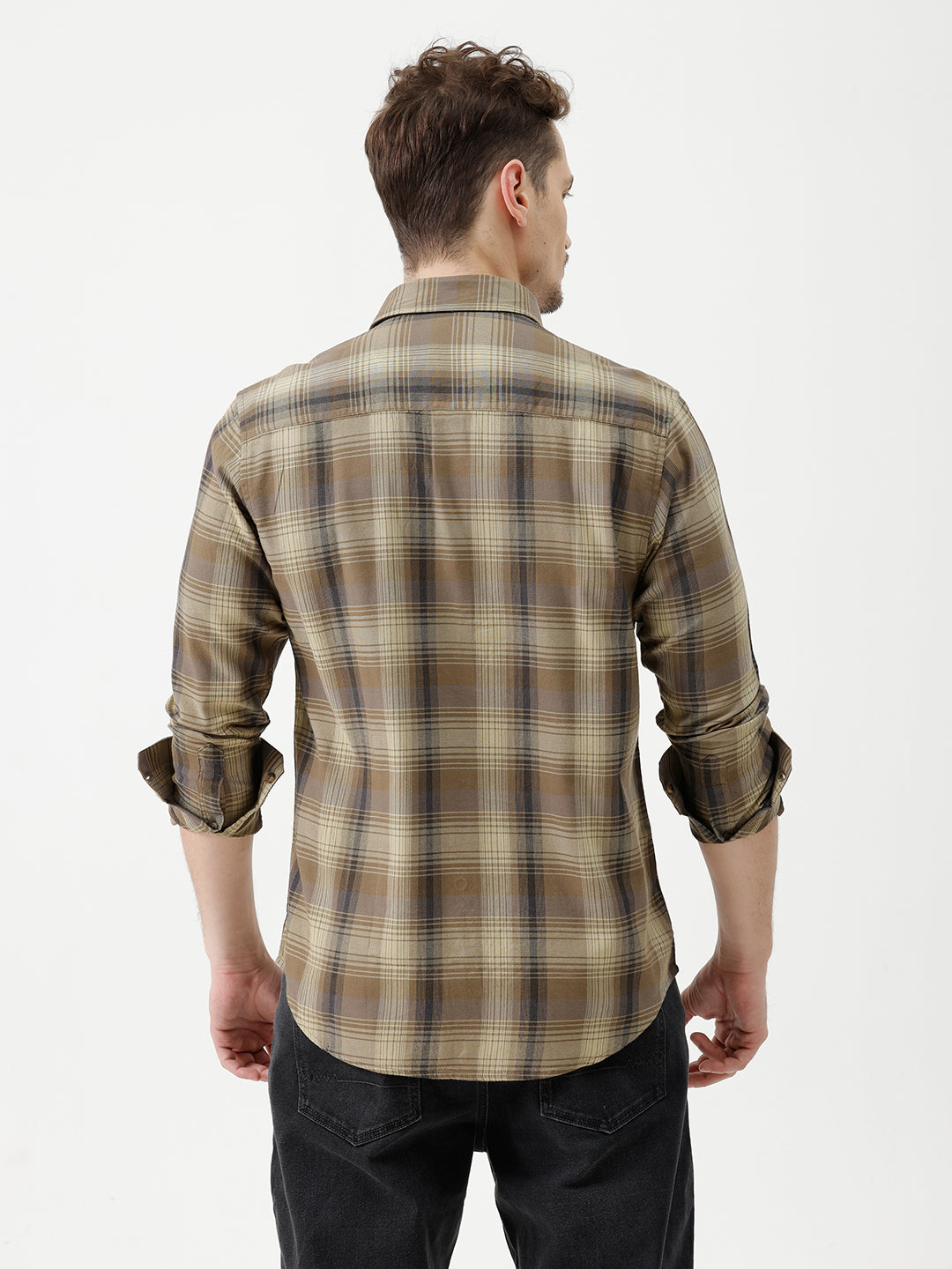 A man wearing a brown and beige plaid long-sleeve button-down shirt with dark sunglasses and black jeans, standing against a plain white studio background.