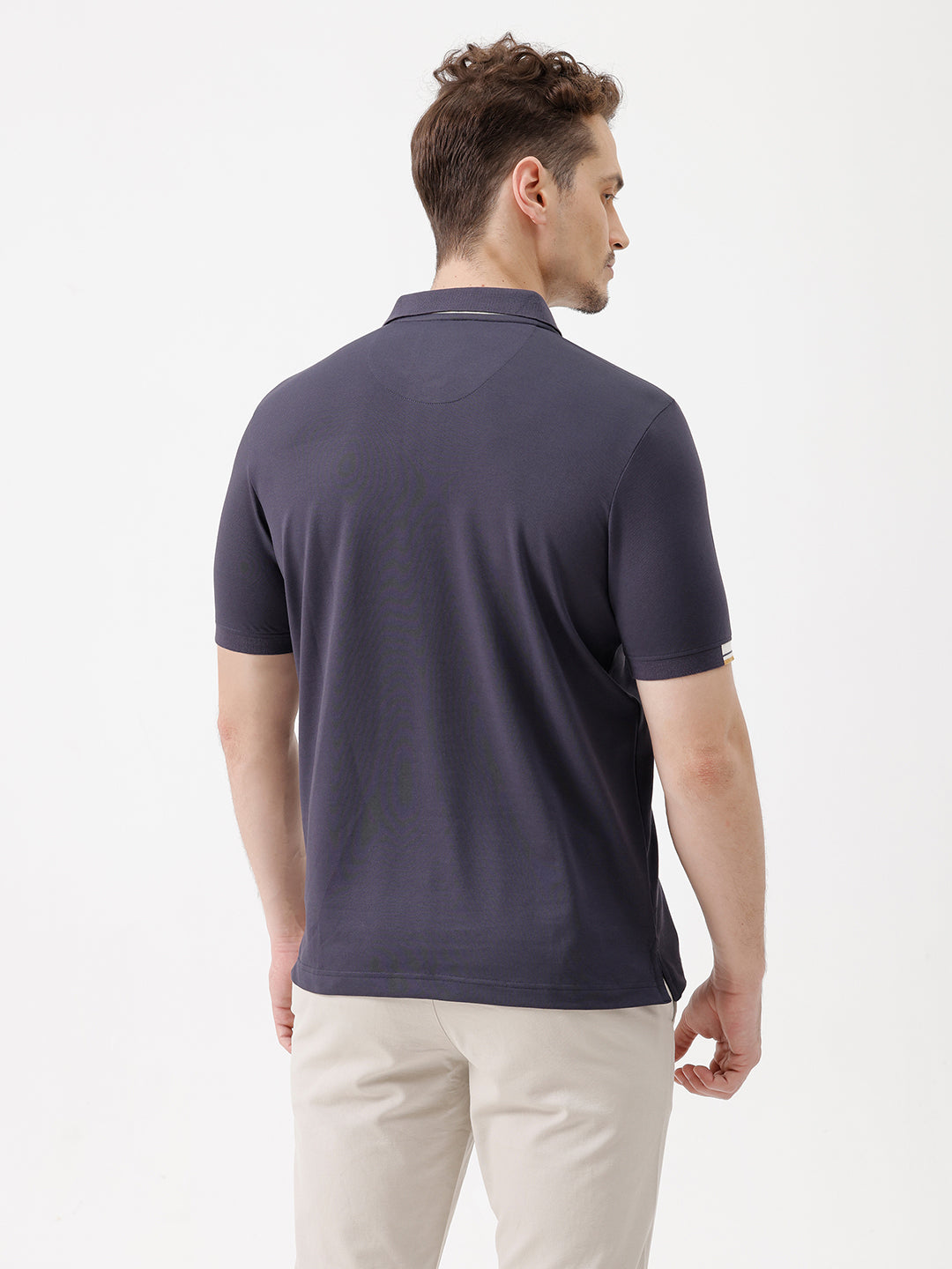 A man wearing a navy blue short-sleeve polo shirt with a contrasting cream and gold striped collar, paired with beige trousers, against a plain white studio background.