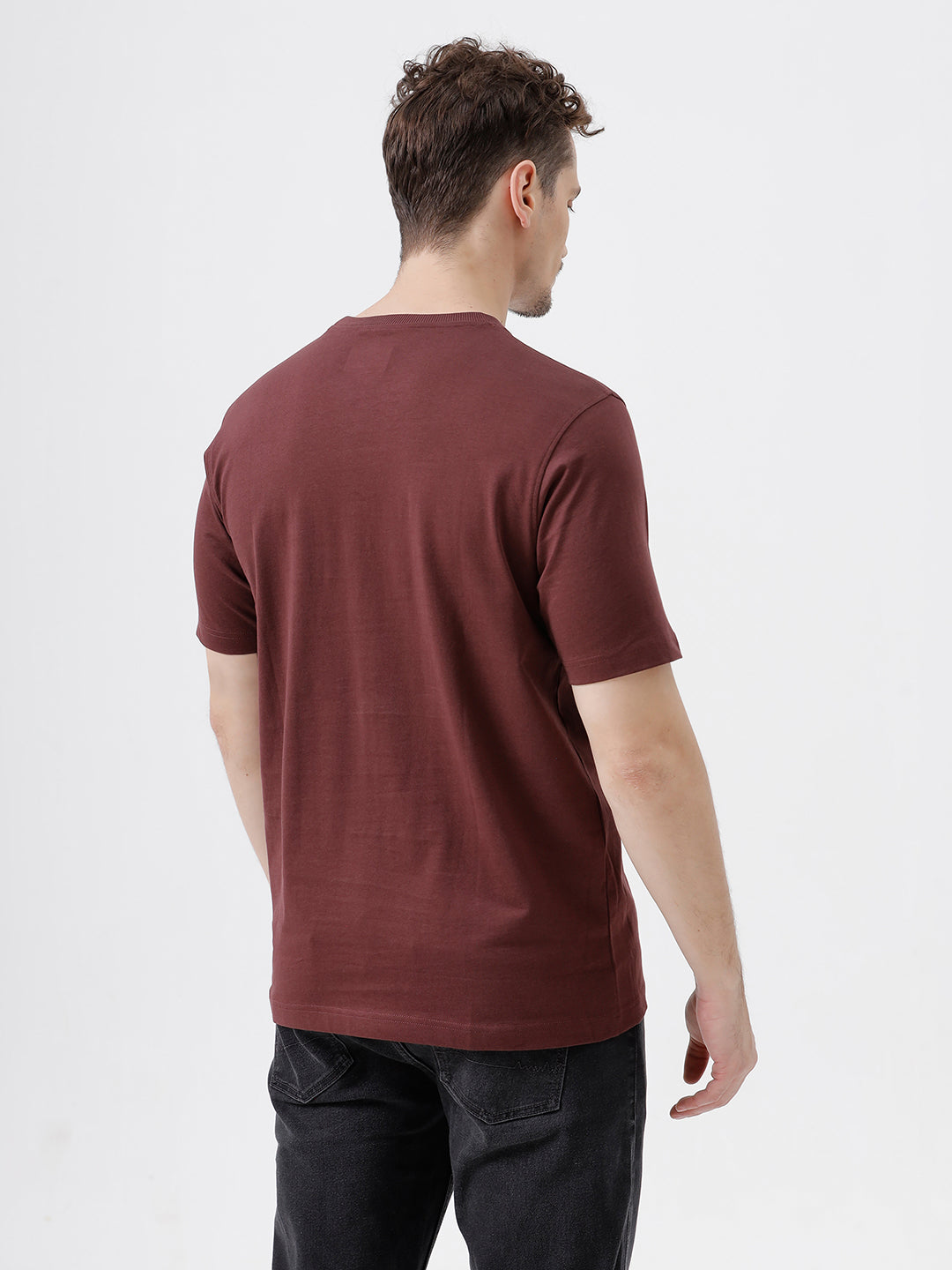 A man wearing a short-sleeve crewneck t-shirt in maroon, paired with dark jeans, standing against a plain light gray studio background.