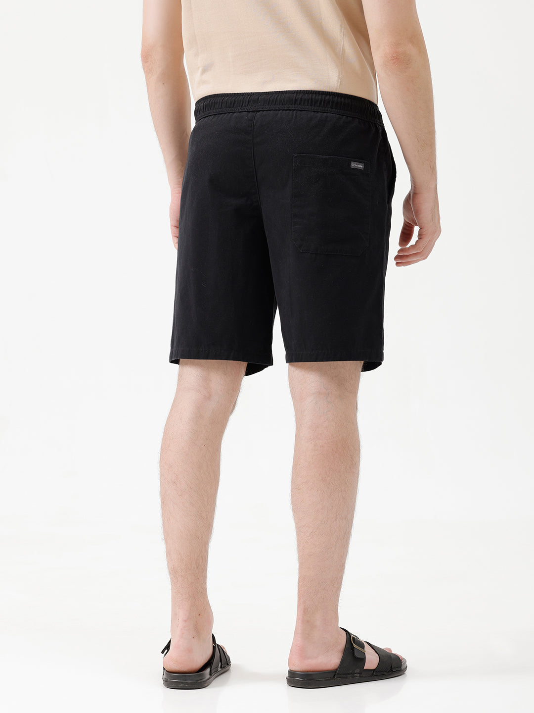 A man modeling a light beige short-sleeve t-shirt, black drawstring shorts, and black slide sandals against a plain white studio background.