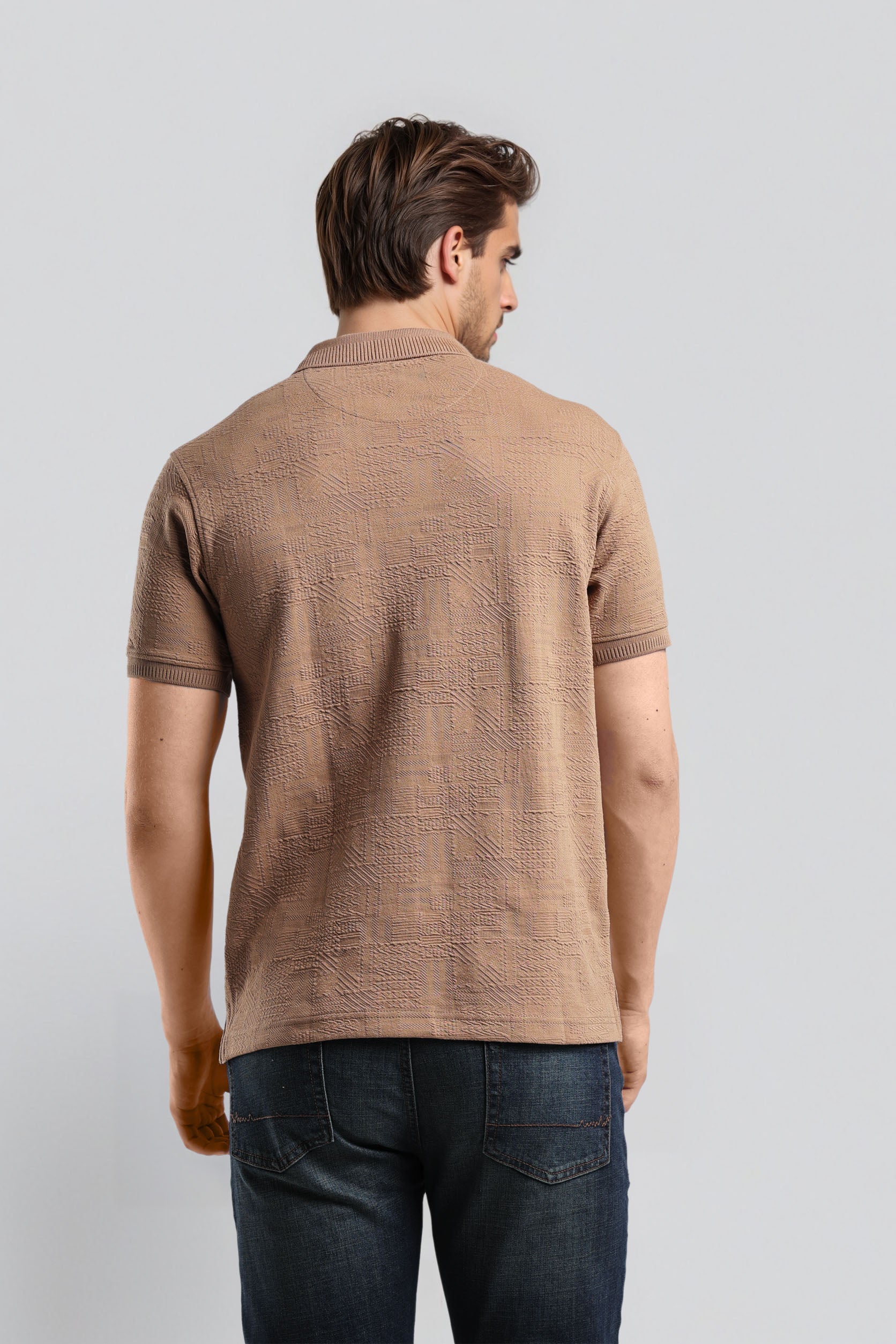 Man wearing a brown patterned polo shirt and dark jeans on a light gray background