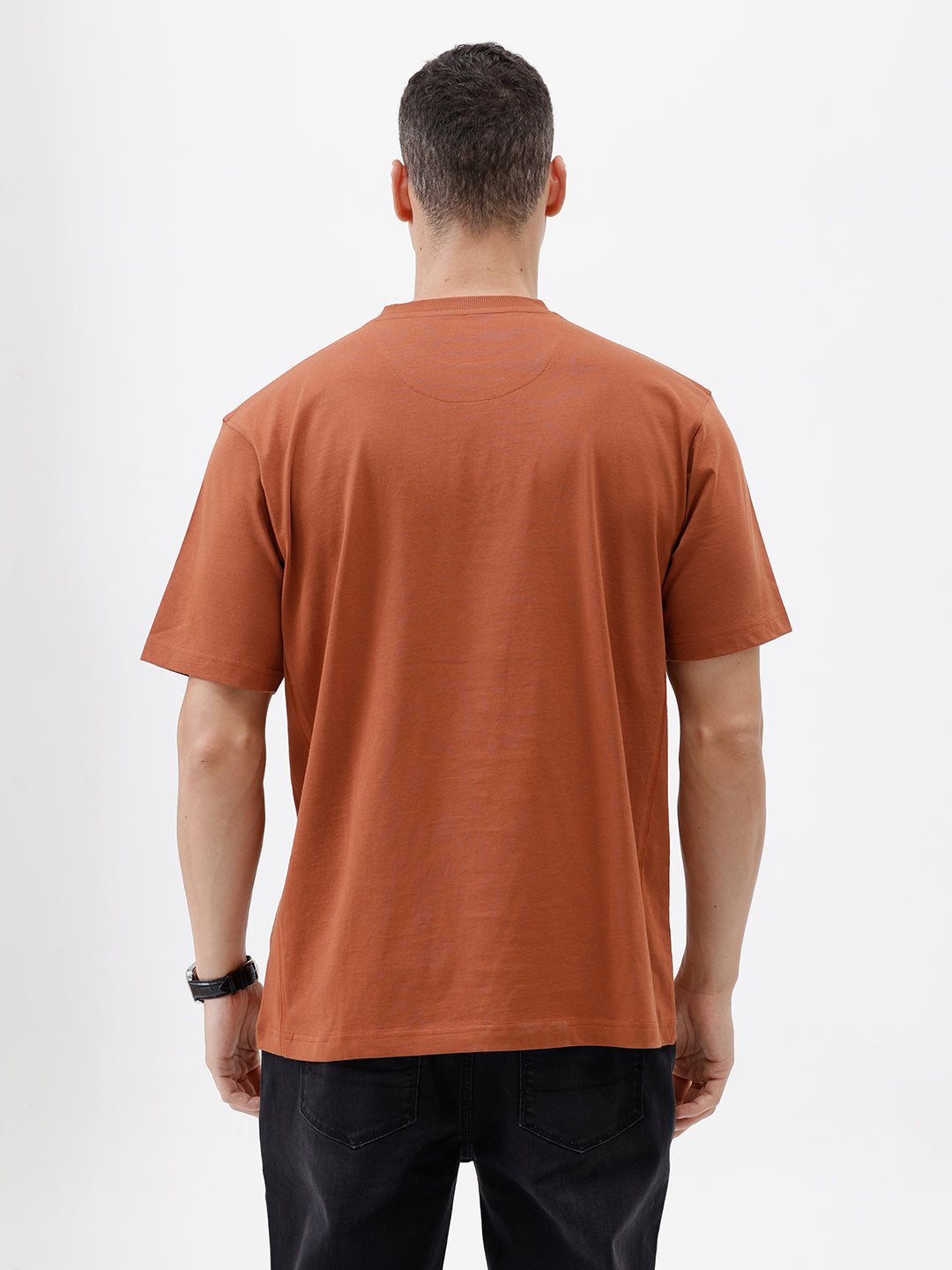 A man wearing a rust-orange crewneck t-shirt with a subtle tonal 'ELECTRIC' logo across the chest, paired with black pants, photographed against a clean white studio background.