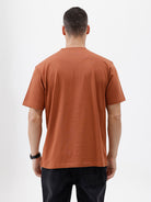 A man wearing a rust-orange crewneck t-shirt with a subtle tonal 'ELECTRIC' logo across the chest, paired with black pants, photographed against a clean white studio background.
