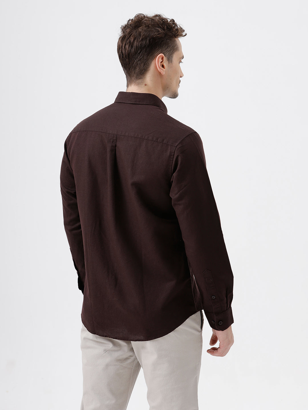 A man wearing a dark brown long-sleeve button-down shirt and light beige trousers, standing against a plain light gray studio background.