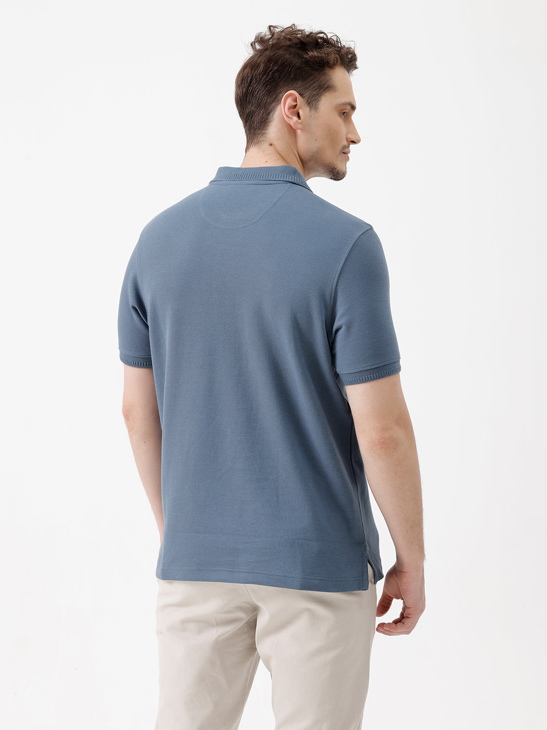 A man wearing a slate blue short-sleeve polo shirt and beige trousers, standing against a plain white studio background.