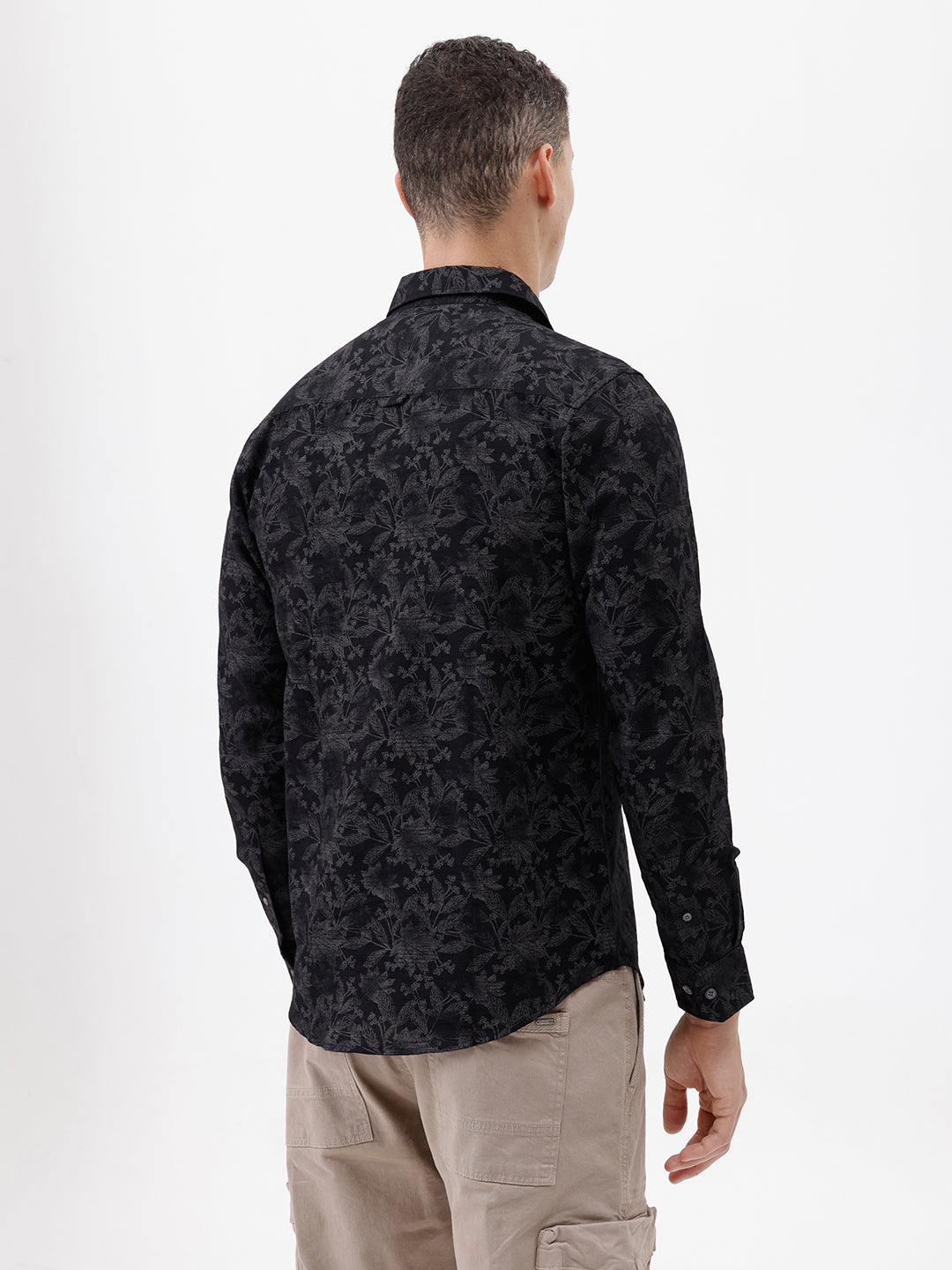 A man wearing a black long-sleeve button-up shirt with a subtle tonal floral pattern, paired with beige cargo pants and dark sunglasses, standing against a plain white background.