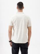 A man wearing a short-sleeve polo shirt in cream with subtle horizontal textured stripes, paired with dark jeans, standing against a plain white studio background.