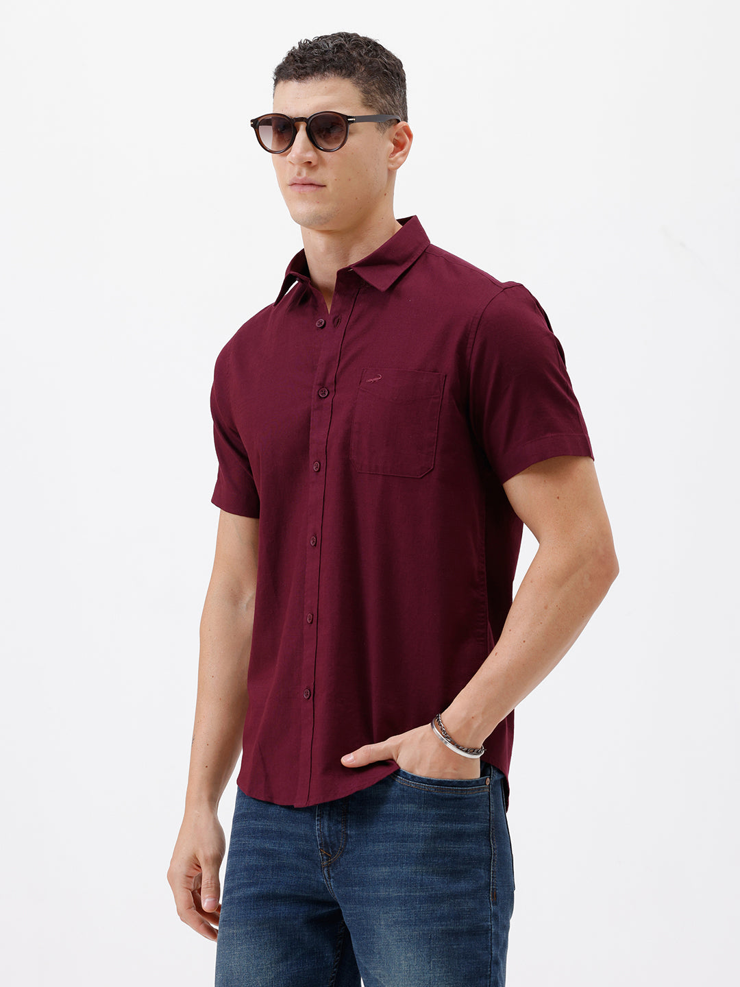 A man wearing a short-sleeve button-up shirt in maroon with a chest pocket, paired with blue jeans, standing against a plain white studio background.