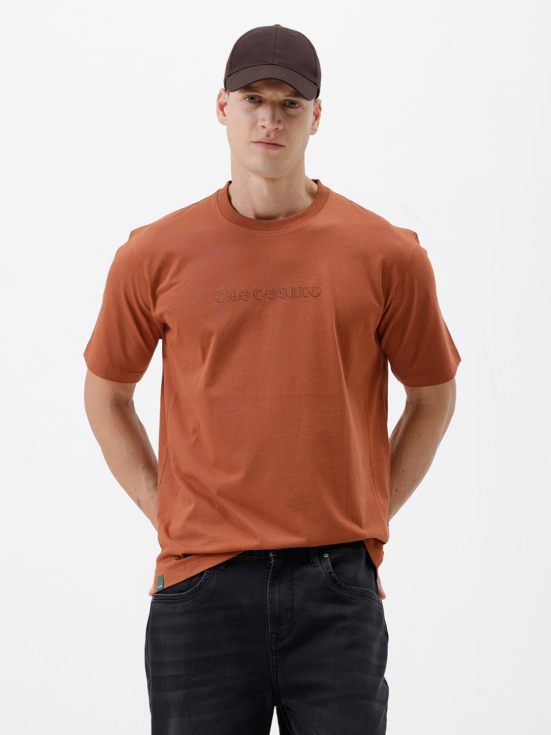 A man wearing a rust-orange crewneck t-shirt with a subtle tonal 'ELECTRIC' logo across the chest, paired with black pants, photographed against a clean white studio background.