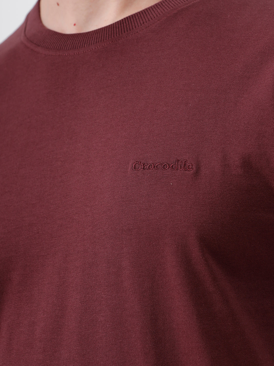 A man wearing a short-sleeve crewneck t-shirt in maroon, paired with dark jeans, standing against a plain light gray studio background.