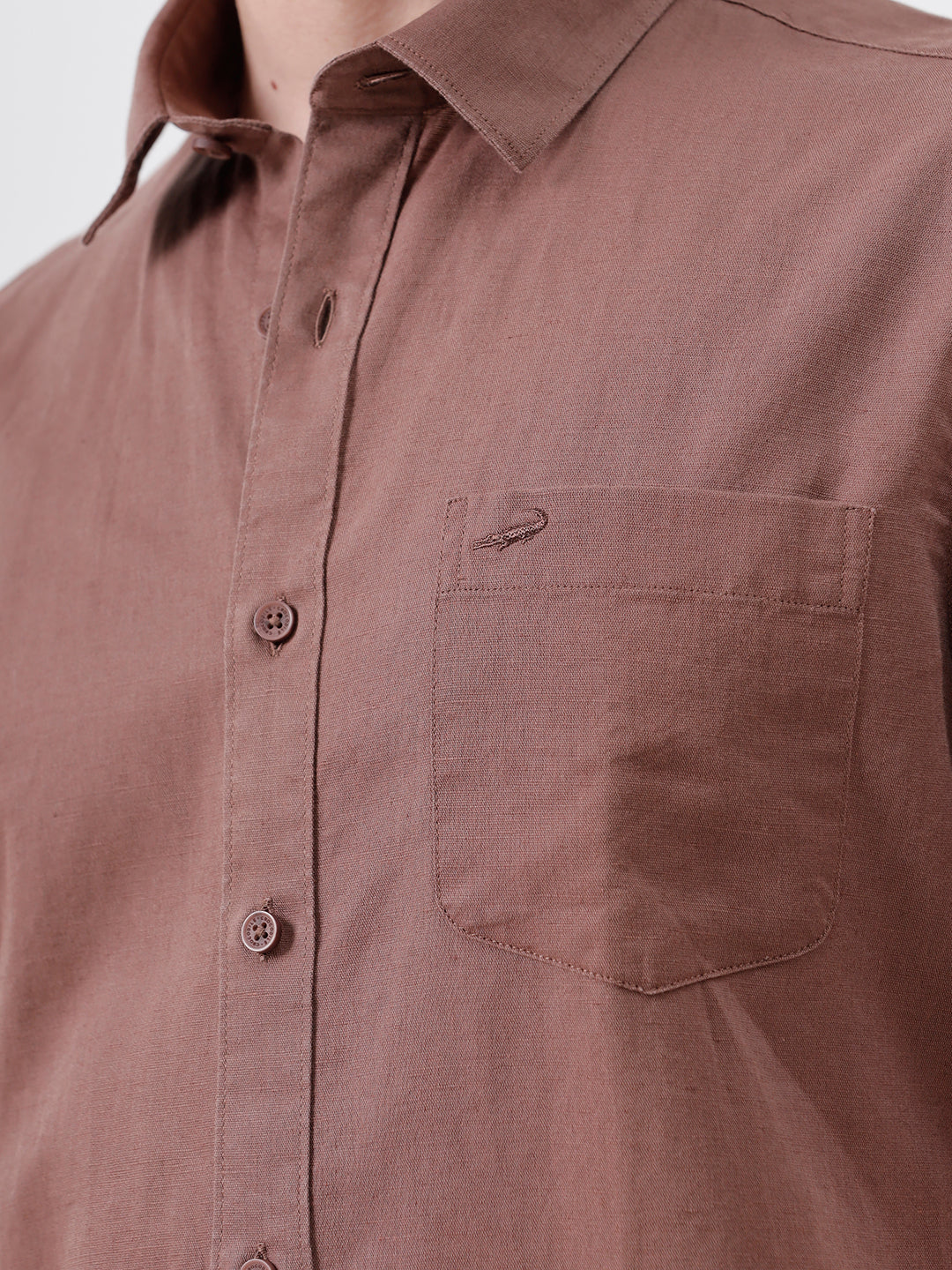 A man wearing a short-sleeve button-up shirt in a dusty rose-brown color, paired with black jeans, standing against a plain light gray background.