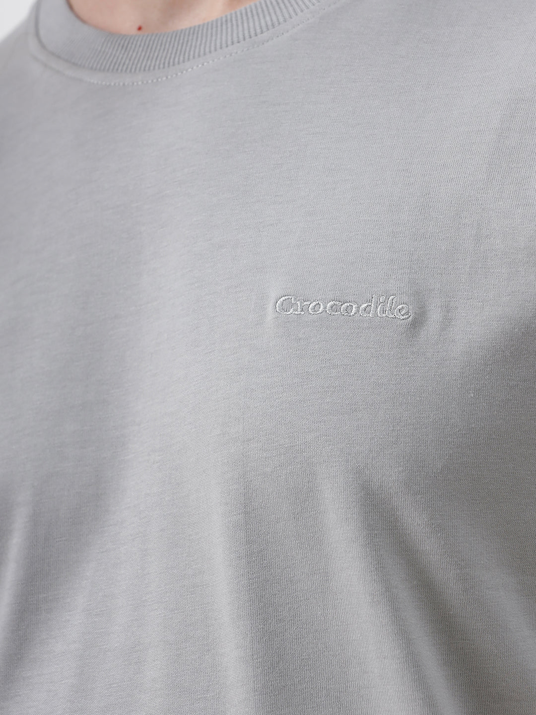 A man wearing a light gray short-sleeve crewneck t-shirt with a subtle tonal chest logo, paired with dark jeans, standing against a light gray studio background.