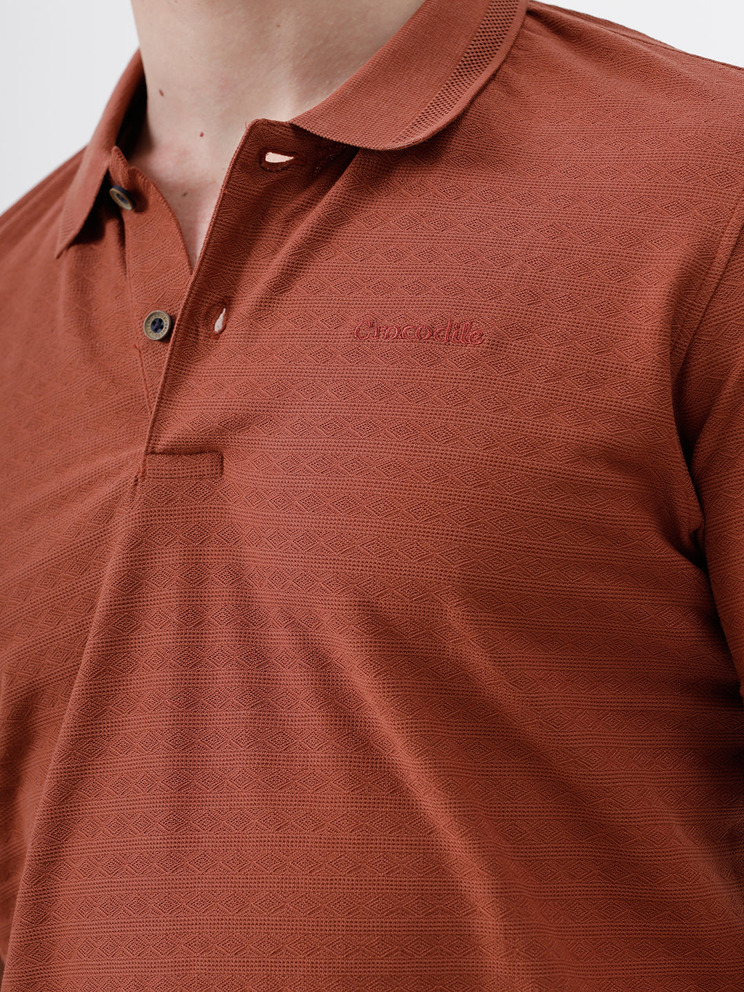 A man wearing a rust-orange short-sleeve polo shirt with a subtle horizontal stripe pattern, paired with dark jeans, standing against a plain white studio background.