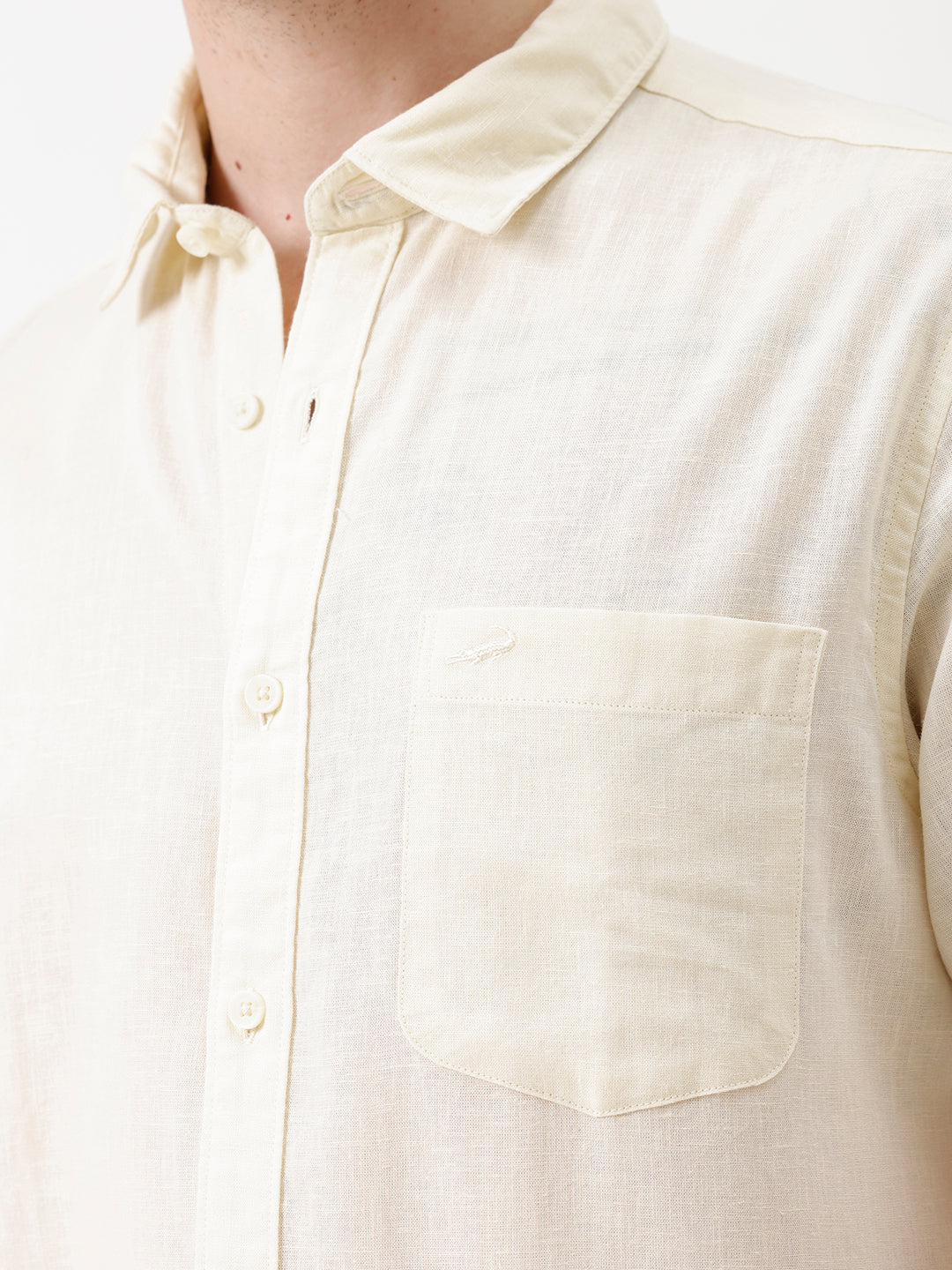 A man wearing a light cream-colored short-sleeve linen button-up shirt with a chest pocket, paired with dark trousers, against a plain white studio background.