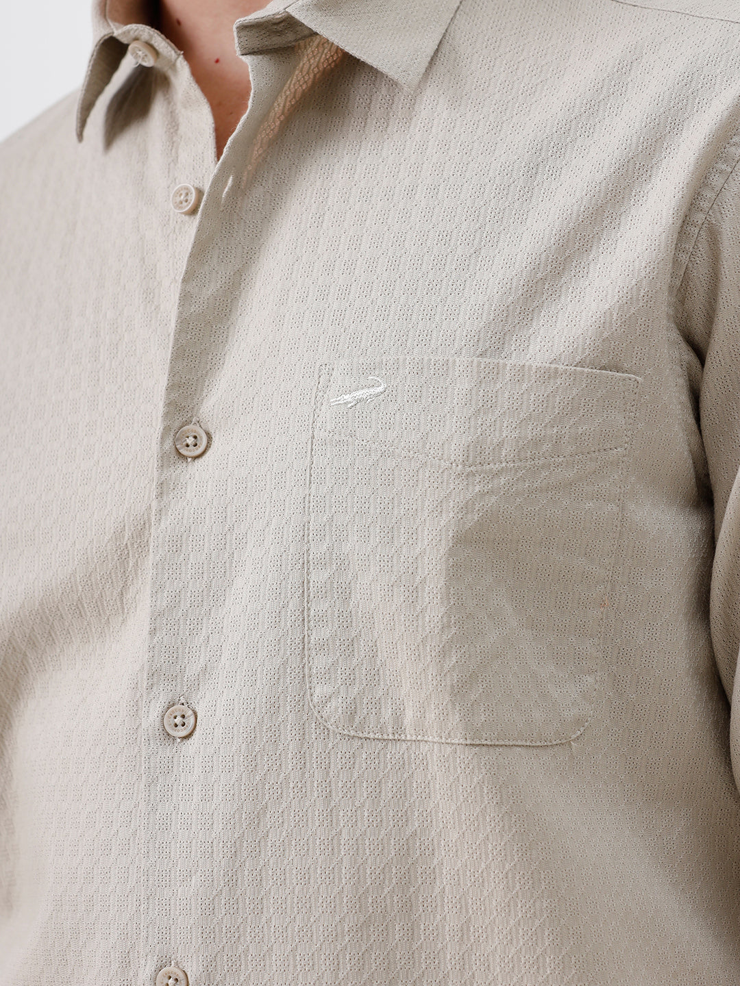 A man wearing a light beige long-sleeve button-down shirt with a chest pocket, paired with dark gray trousers, standing against a plain white studio background.