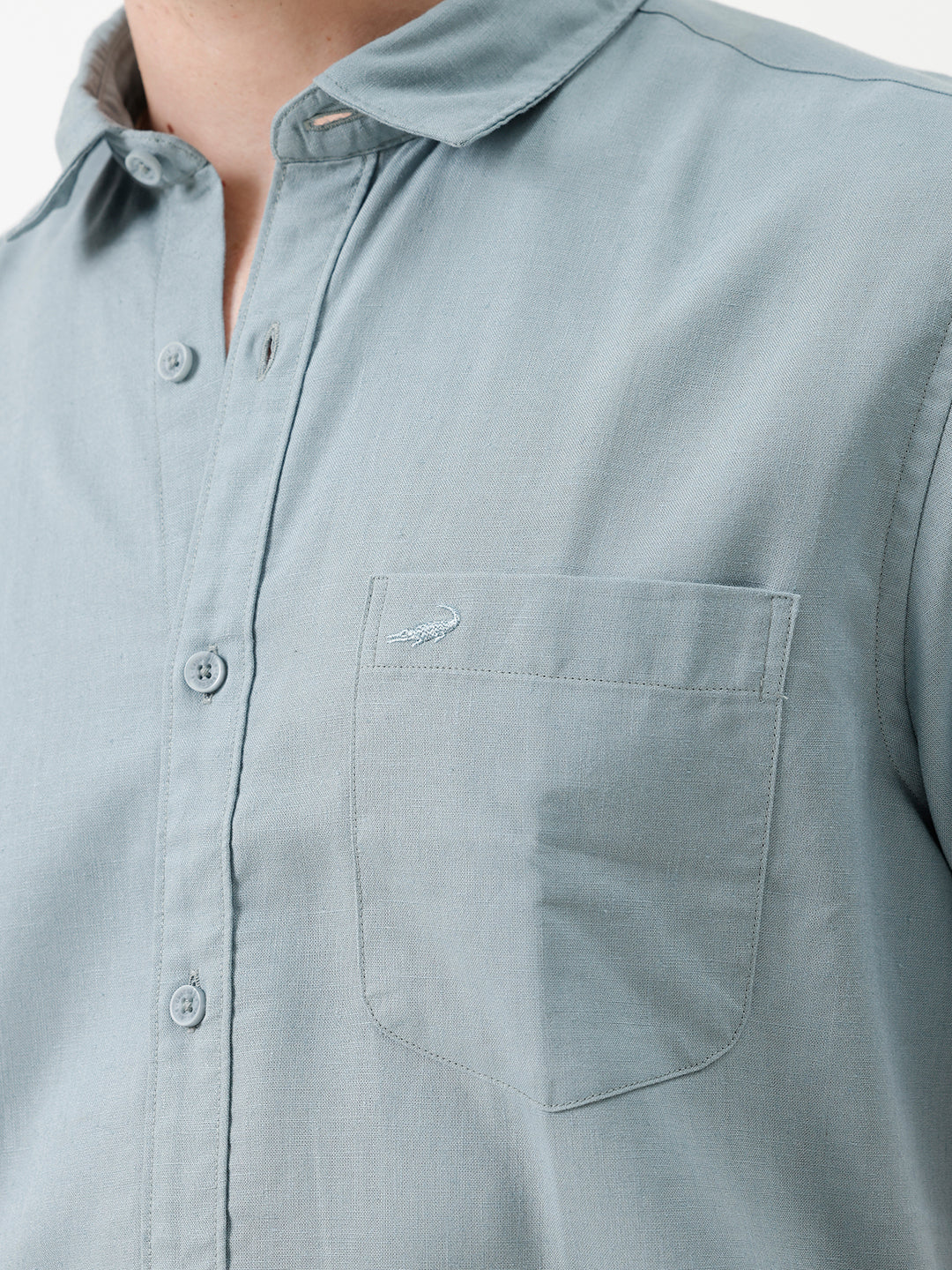 A man wearing a light blue short-sleeve button-up shirt and beige trousers, standing against a plain white studio background.