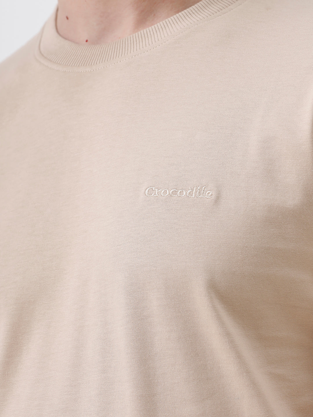 A man wearing a beige short-sleeve crewneck t-shirt with a subtle tonal logo on the left chest, paired with black jeans, standing against a light gray studio background.
