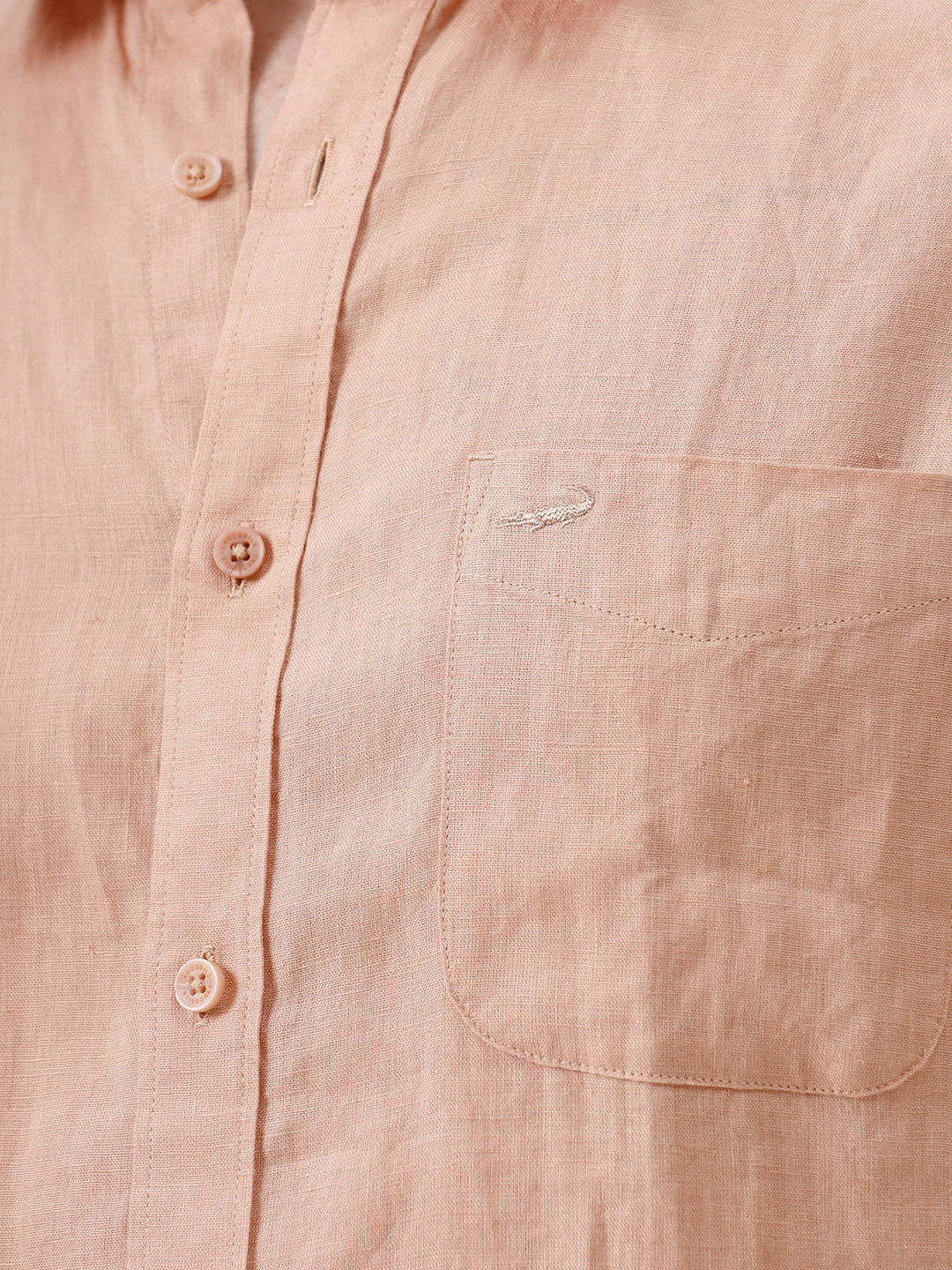 A man wearing a short-sleeve linen button-up shirt in a light peach-pink color, paired with light gray trousers, standing against a plain light gray background.