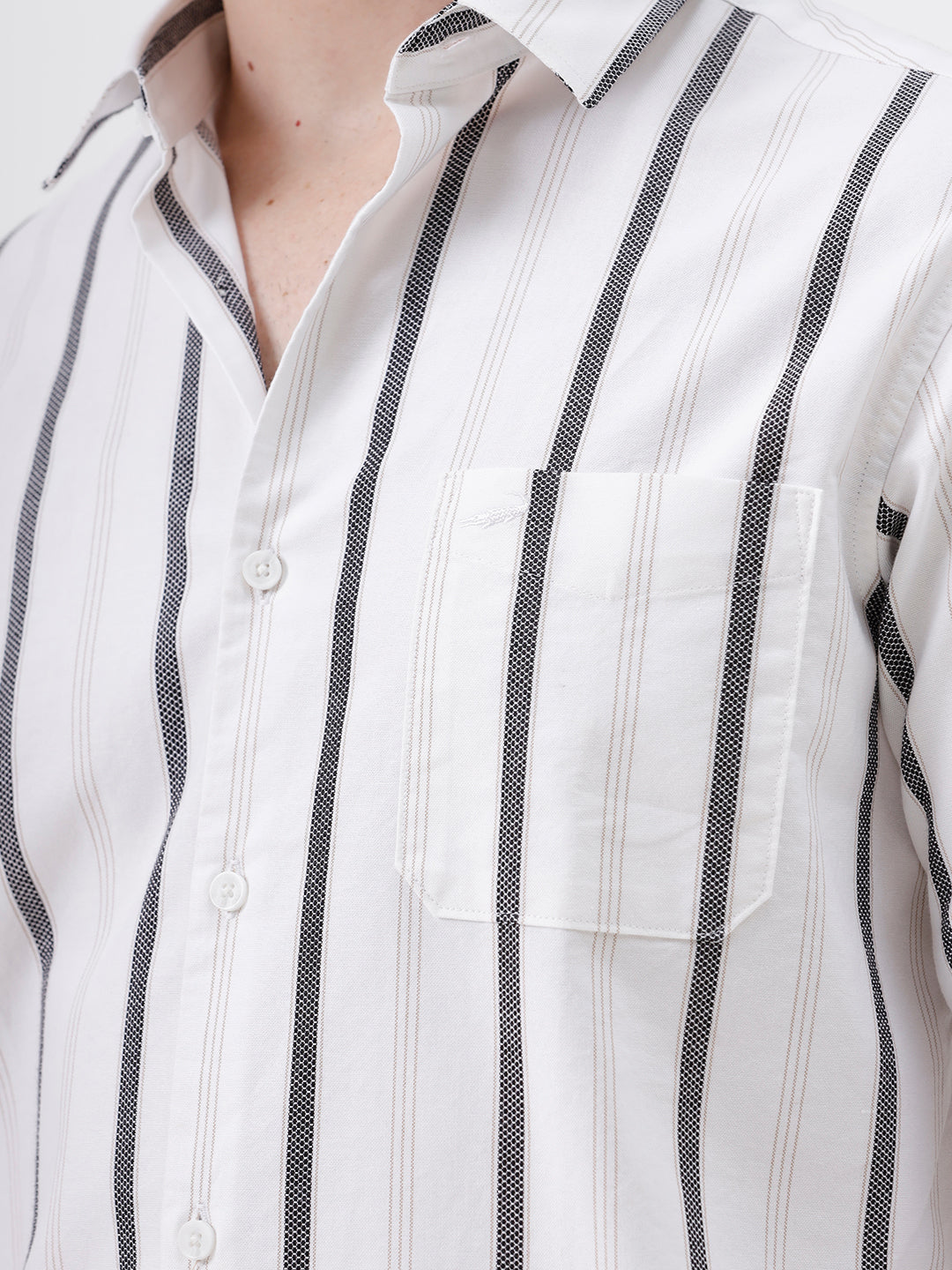 A man wearing a white long-sleeve button-up shirt with vertical gray stripes and beige trousers, standing against a light gray studio background.