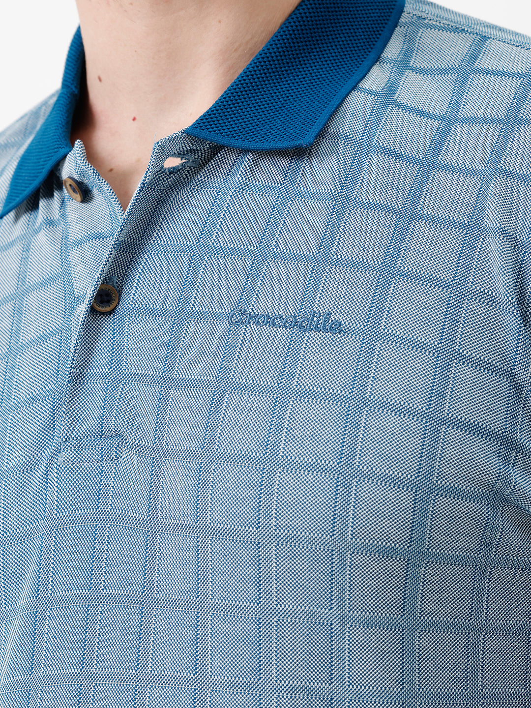 A man wearing a light blue and white checkered polo shirt with a dark blue collar and sleeve cuffs, paired with white trousers, standing against a plain white background.