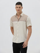 A man wearing a short-sleeve linen button-up shirt with a color-blocked design in beige and cream, paired with black pants, against a plain white studio background.