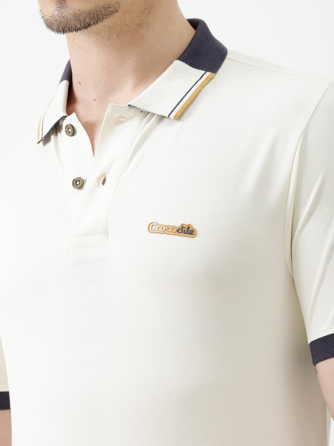 A man wearing a short-sleeve polo shirt in off-white with navy blue and gold trim on the collar and sleeve cuffs, paired with dark navy trousers, against a plain light gray background.