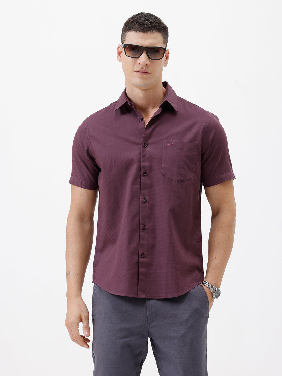 A man modeling a short-sleeve button-up shirt in a deep plum purple color, paired with gray trousers, against a plain white studio background.