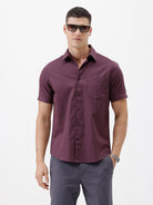 A man modeling a short-sleeve button-up shirt in a deep plum purple color, paired with gray trousers, against a plain white studio background.