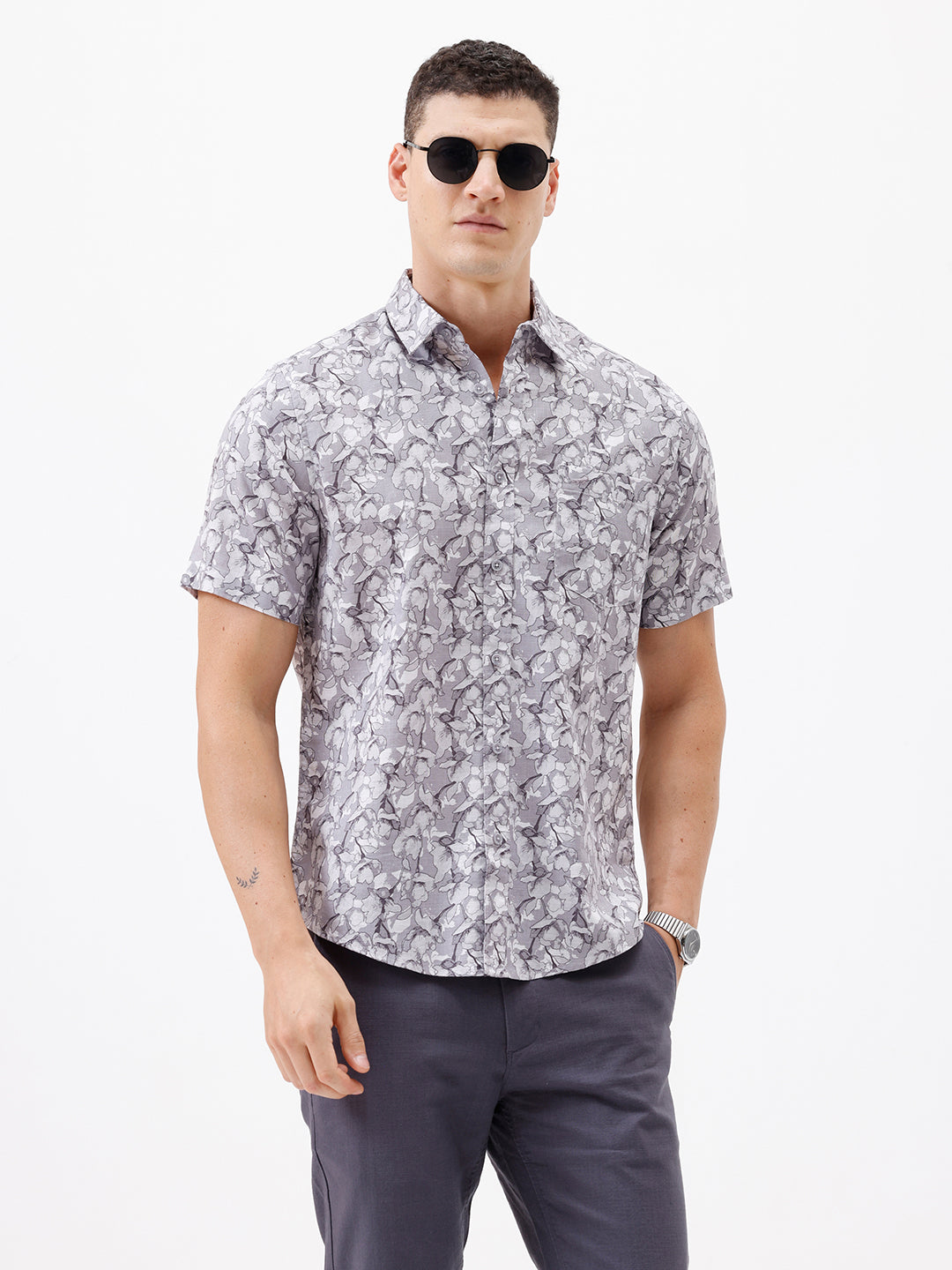A man wearing a short-sleeve button-up shirt with a gray and white abstract pattern, paired with dark gray trousers, standing against a plain white studio background.