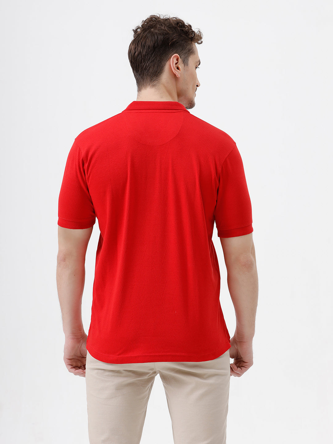 A man wearing a bright red short-sleeve polo shirt with a chest pocket, paired with beige trousers, standing against a plain white studio background.